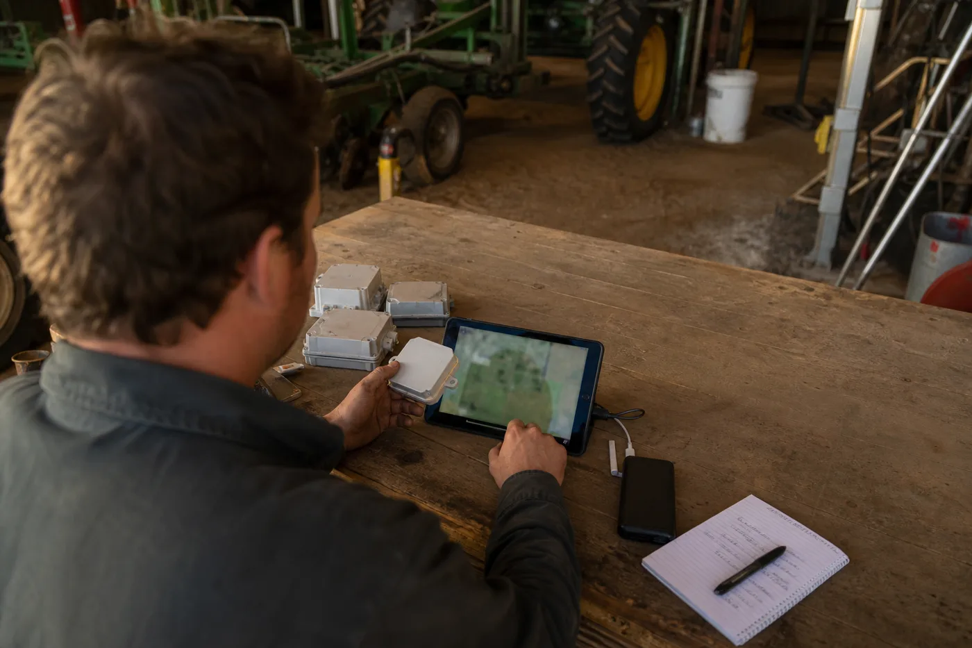A person holding a small white box and touching a tablet on a wooden table in a barn.