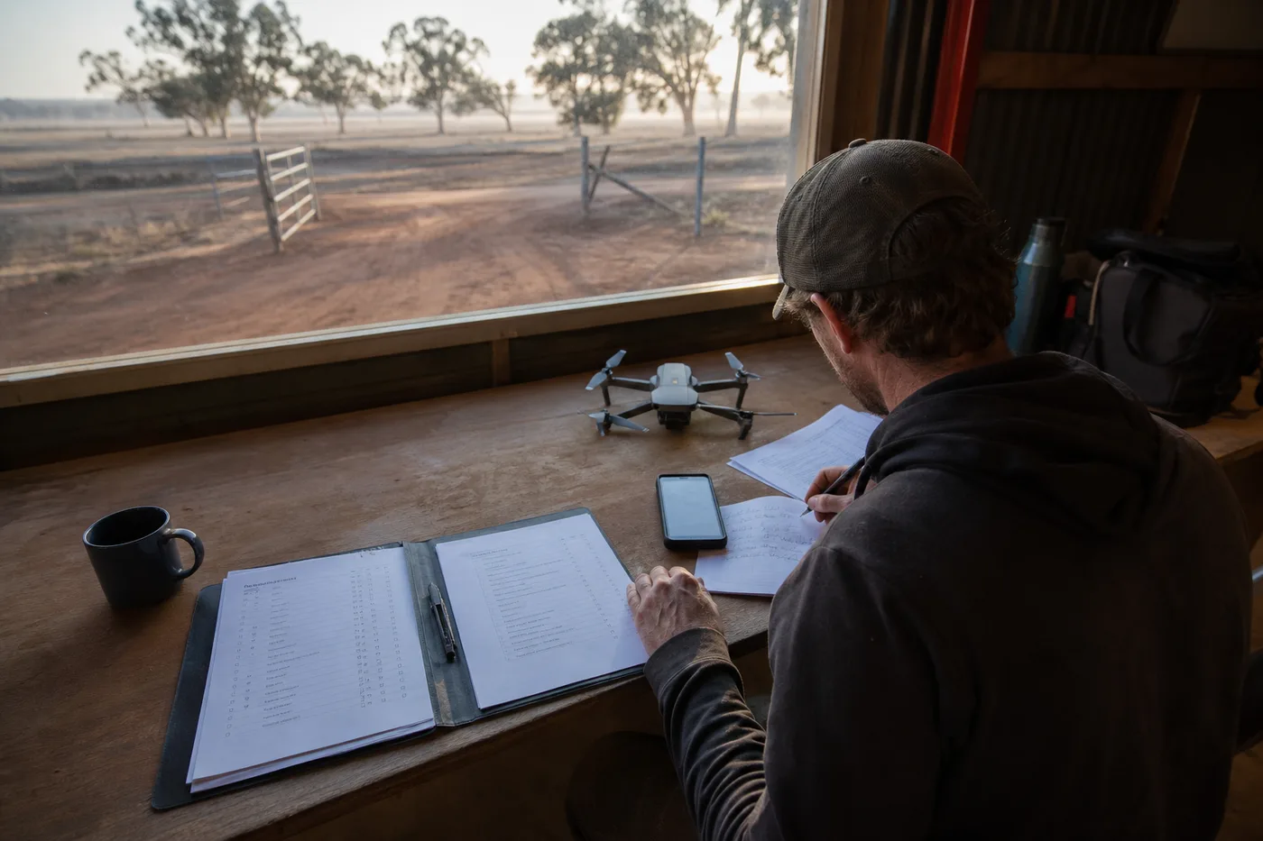 A person sits at a wooden desk with papers, a phone, and a small drone, looking out a large window at a dirt field.