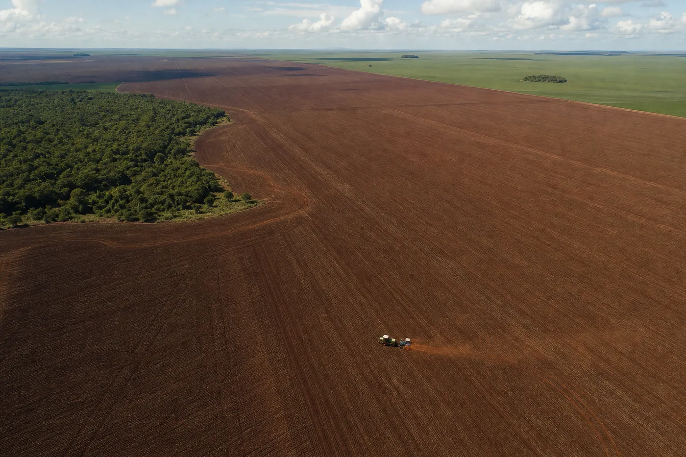 An aerial view of a tractor tilling a vast brown field bordered by green forest and distant plains.
