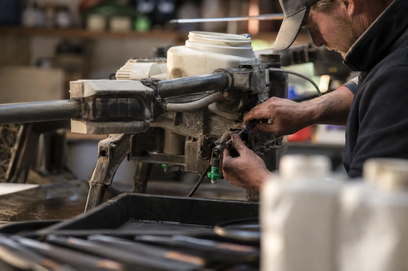 A person wearing a cap holds a tool and works on a complex machine with tubes and a white tank.