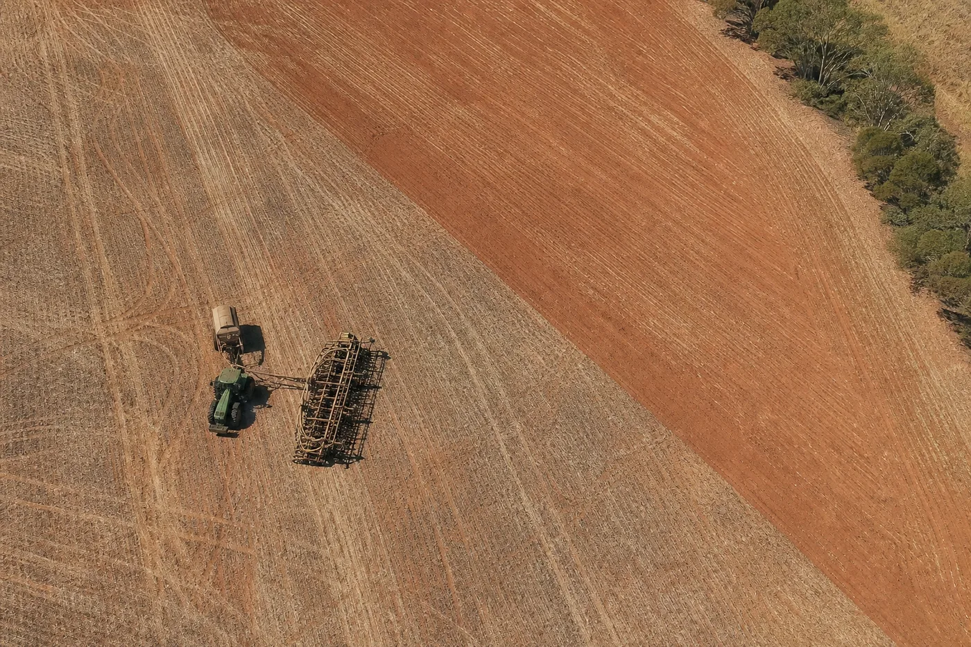 An aerial view of a tractor pulling heavy equipment across a divided field of red and dry brown soil.