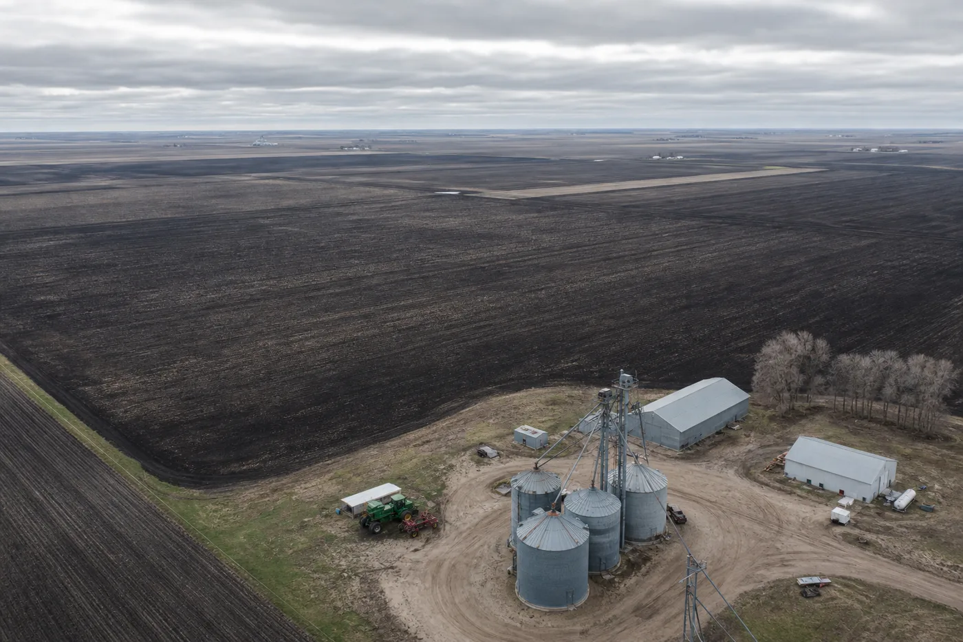 An aerial view of flat, dark farmland stretching to a distant horizon under a cloudy sky, with a homestead below.