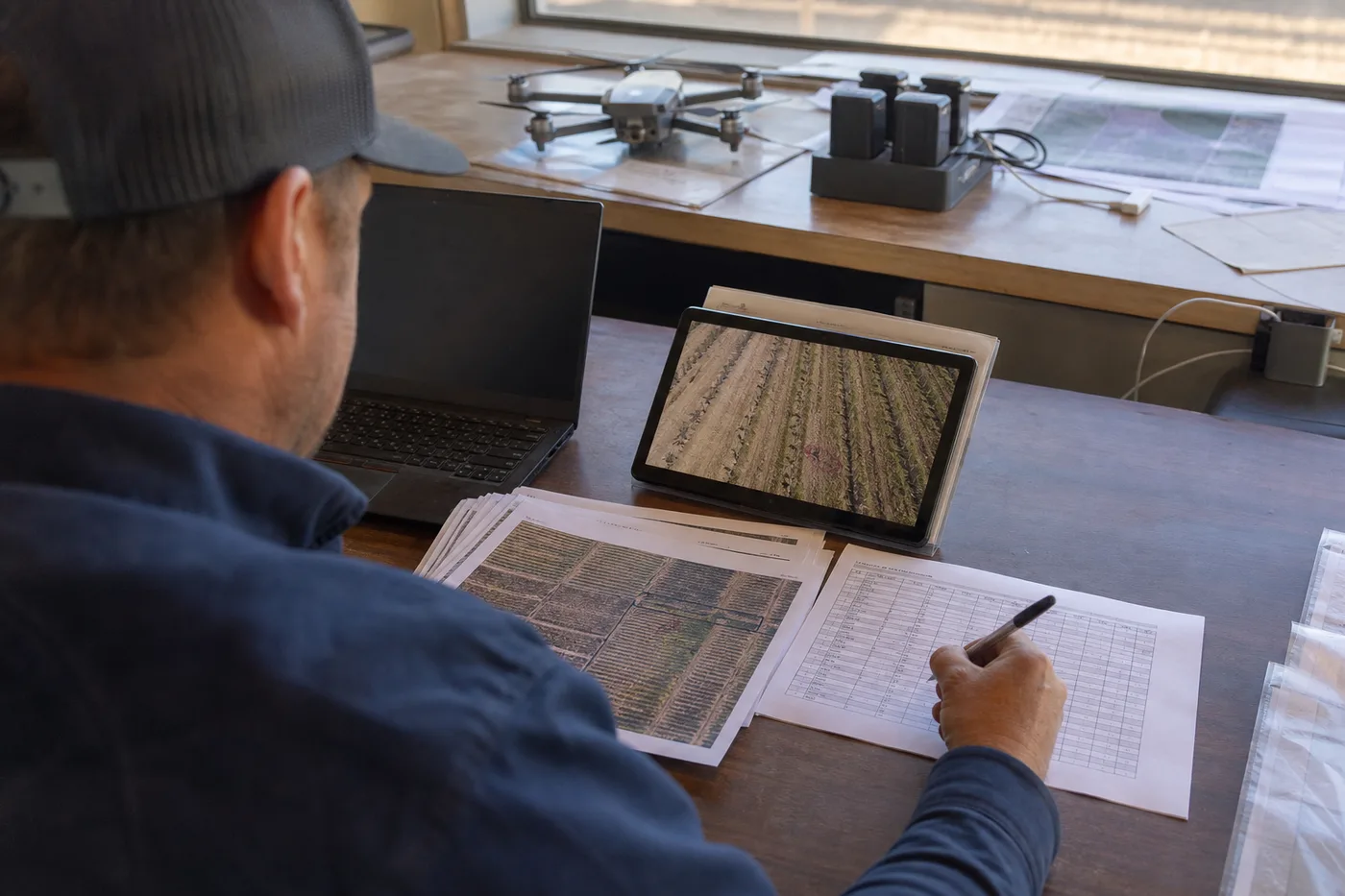 A person wearing a cap reviews documents and a tablet displaying aerial field images at a desk.