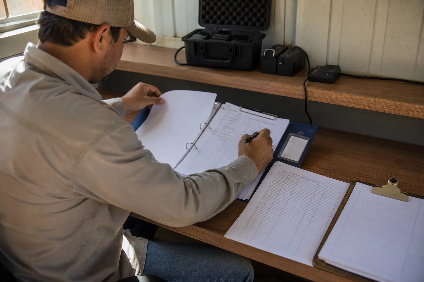 A person wearing a baseball cap sits at a wooden desk, holding a pen and turning a page in a binder.
