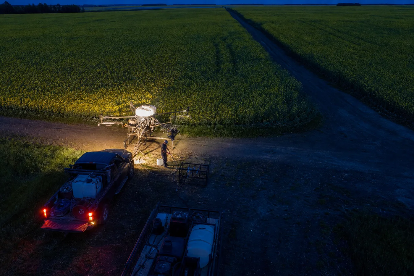 A person works near a pickup truck and a drone on a dirt road beside a large field at dusk.
