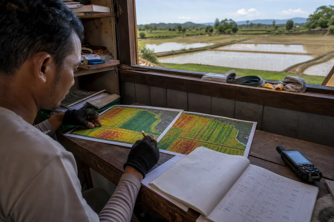A person in gloves sits indoors at a desk, reviewing color-coded maps alongside a notebook and an open window showing fields.