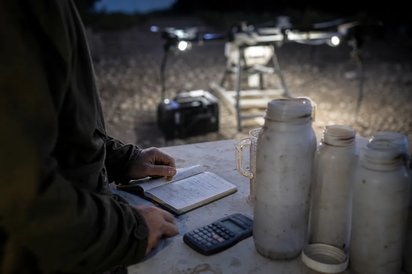 A person writes in a notebook on a table beside plastic containers, with a glowing drone in the background.