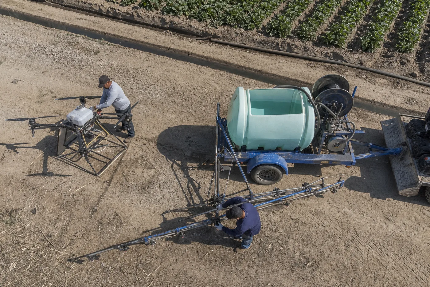 Two workers prep an agricultural drone near a large water tank trailer next to a plowed field.