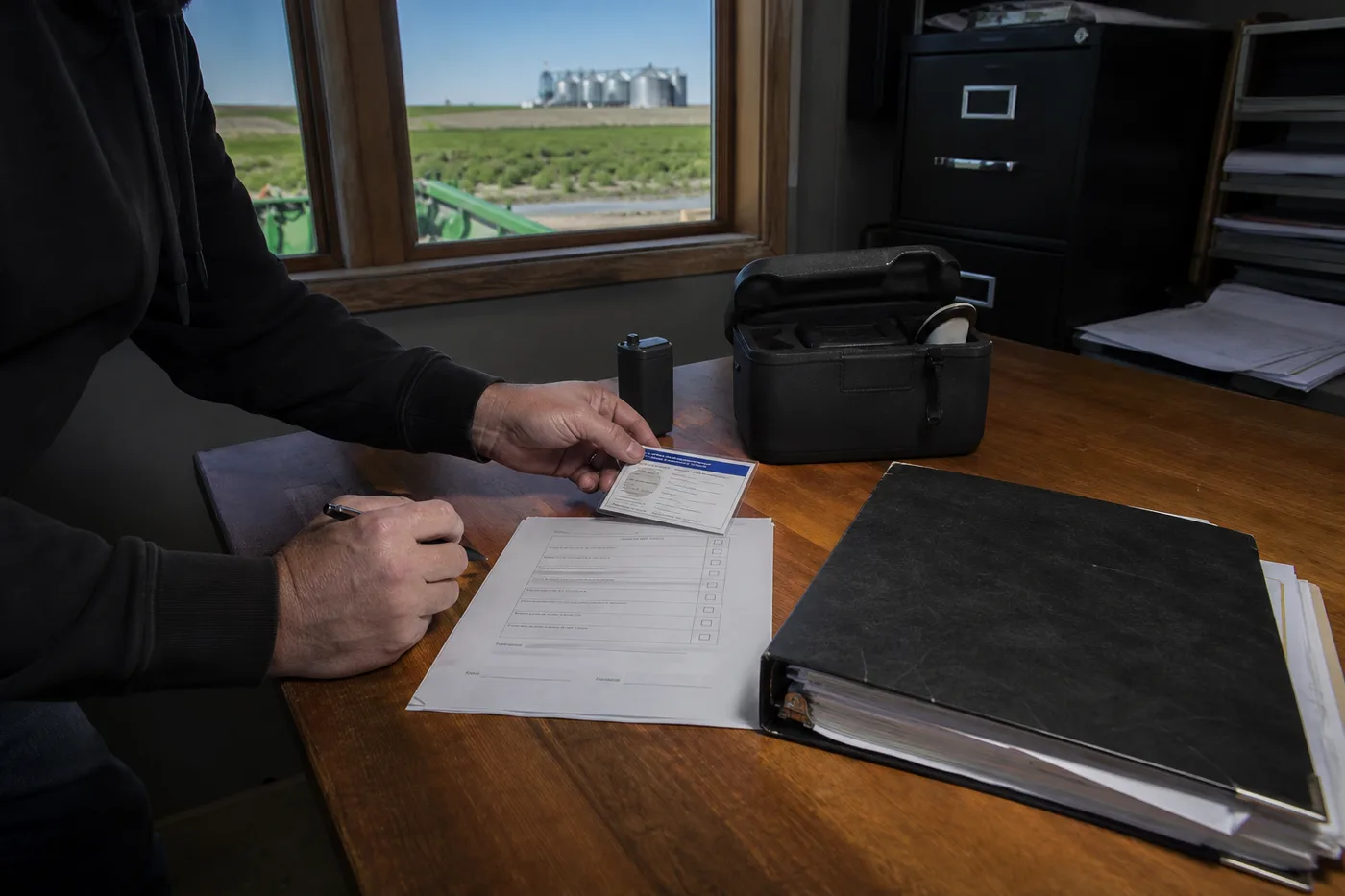 A person in a dark hoodie holds a card over a paper form on a wooden desk.
